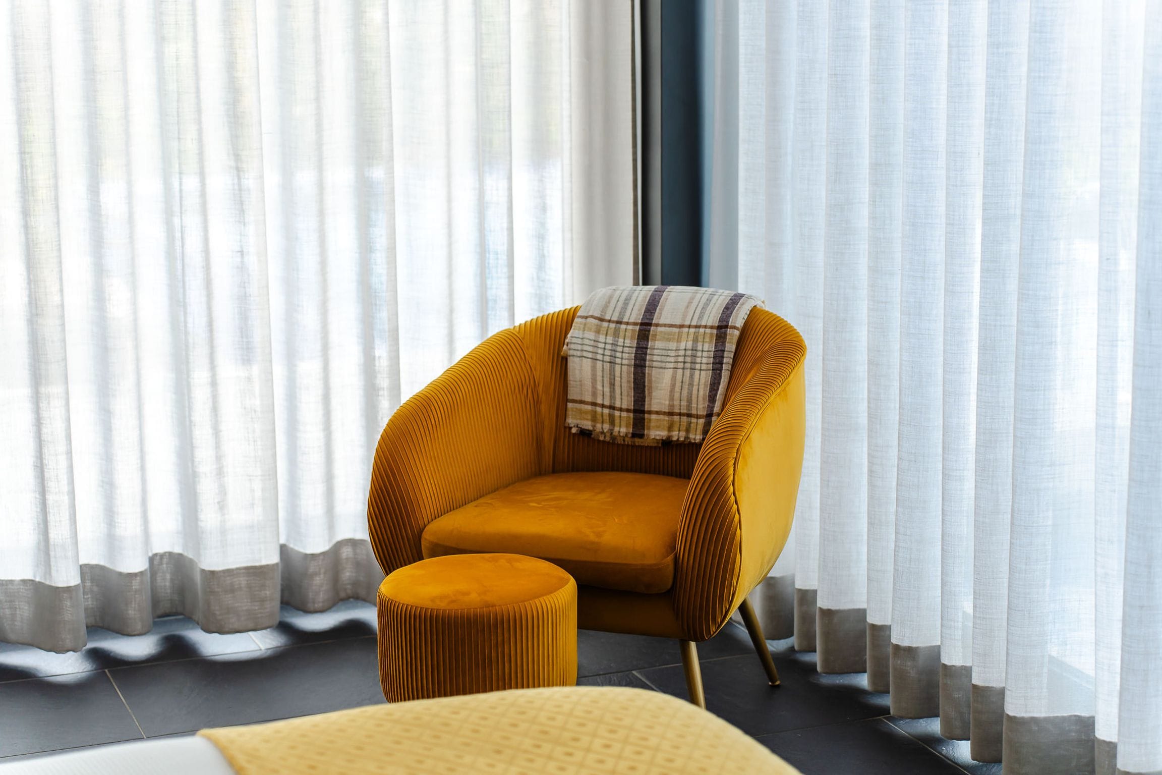 Ochre velvet armchair and footstool in front of sheer curtains