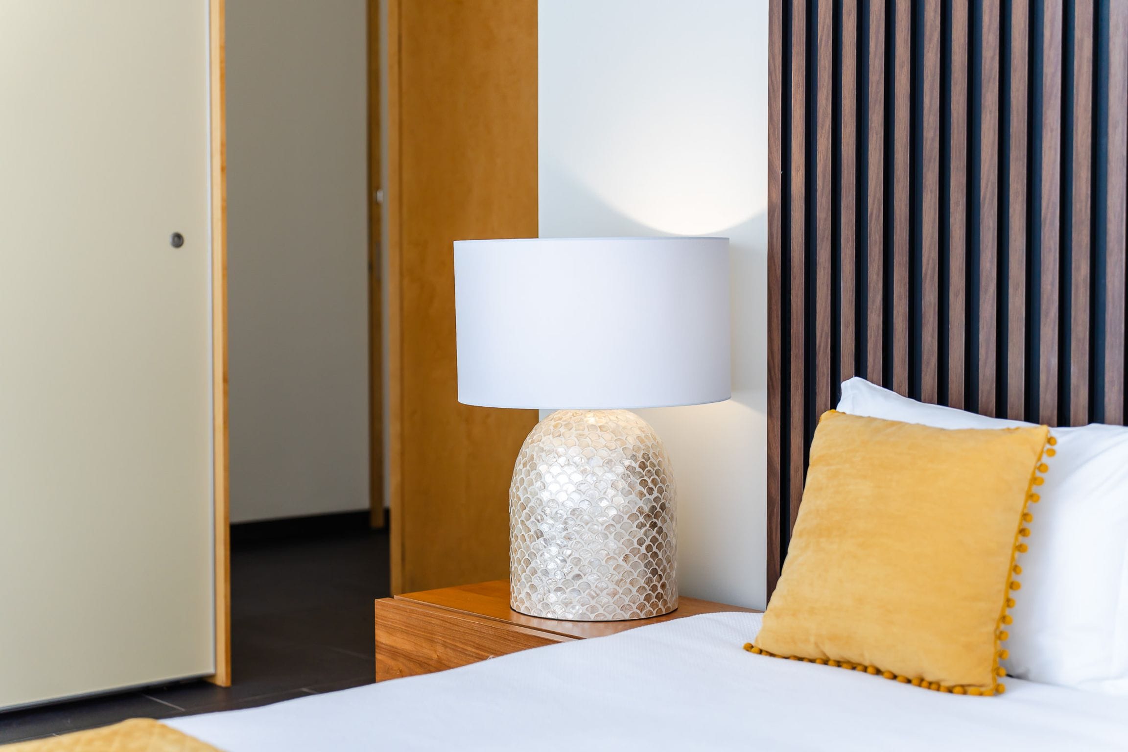 Mother-of-pearl bedside lamp and ochre cushion against a slatted timber wall