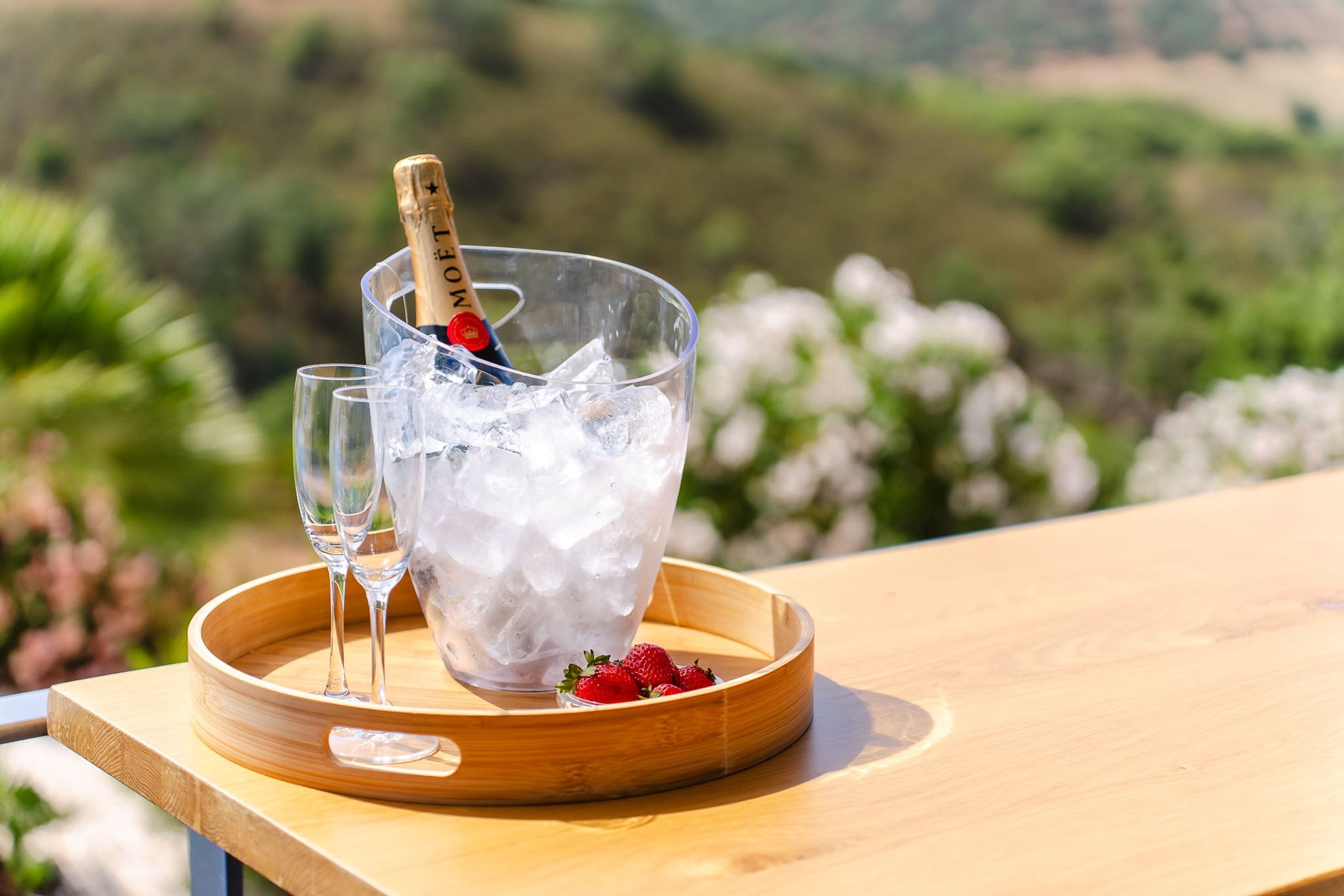 Champagne on ice with two glasses and strawberries on a timber tray, hills softly blurred behind