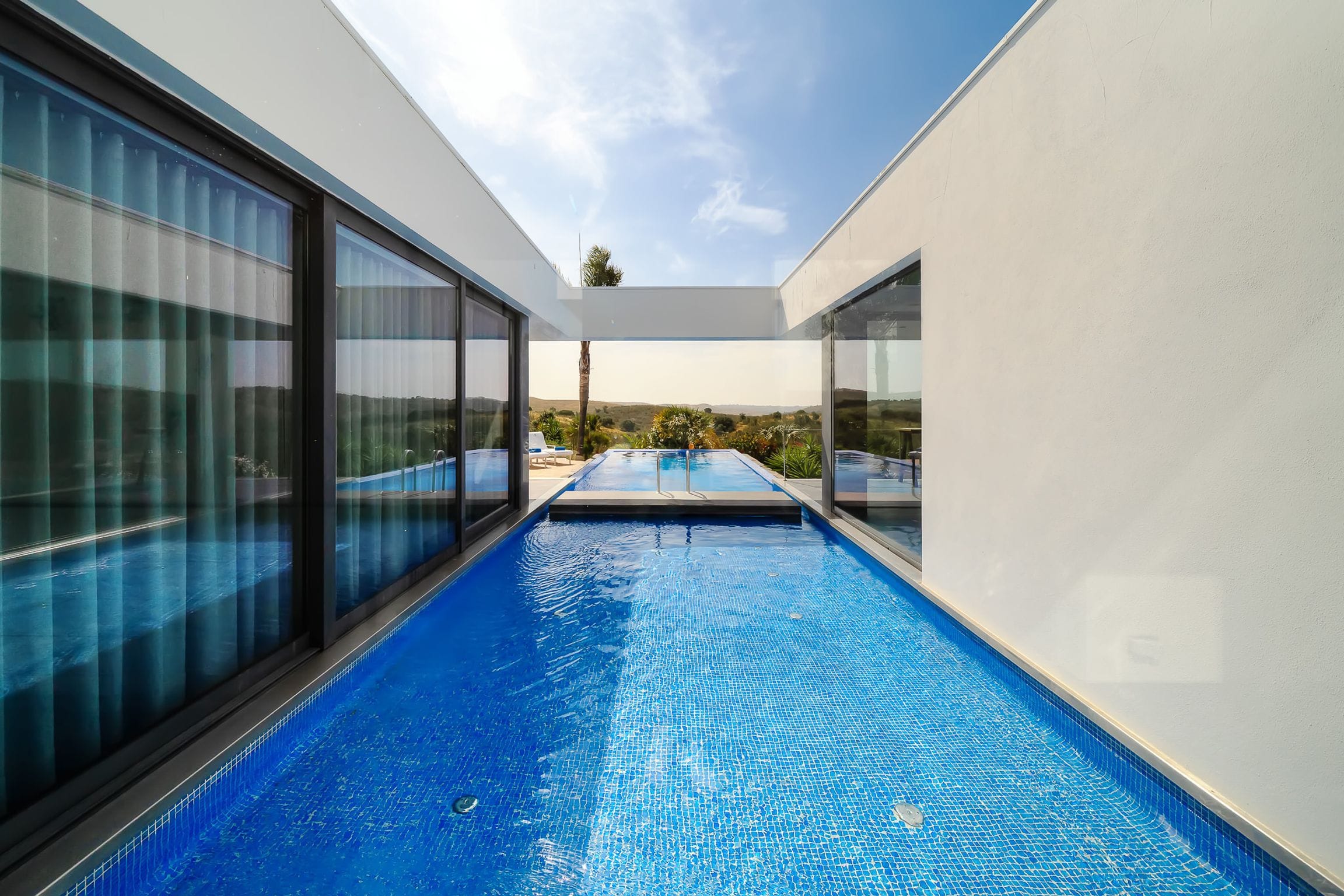 Narrow channel of pool water running between floor-to-ceiling glass and a white wall towards the landscape