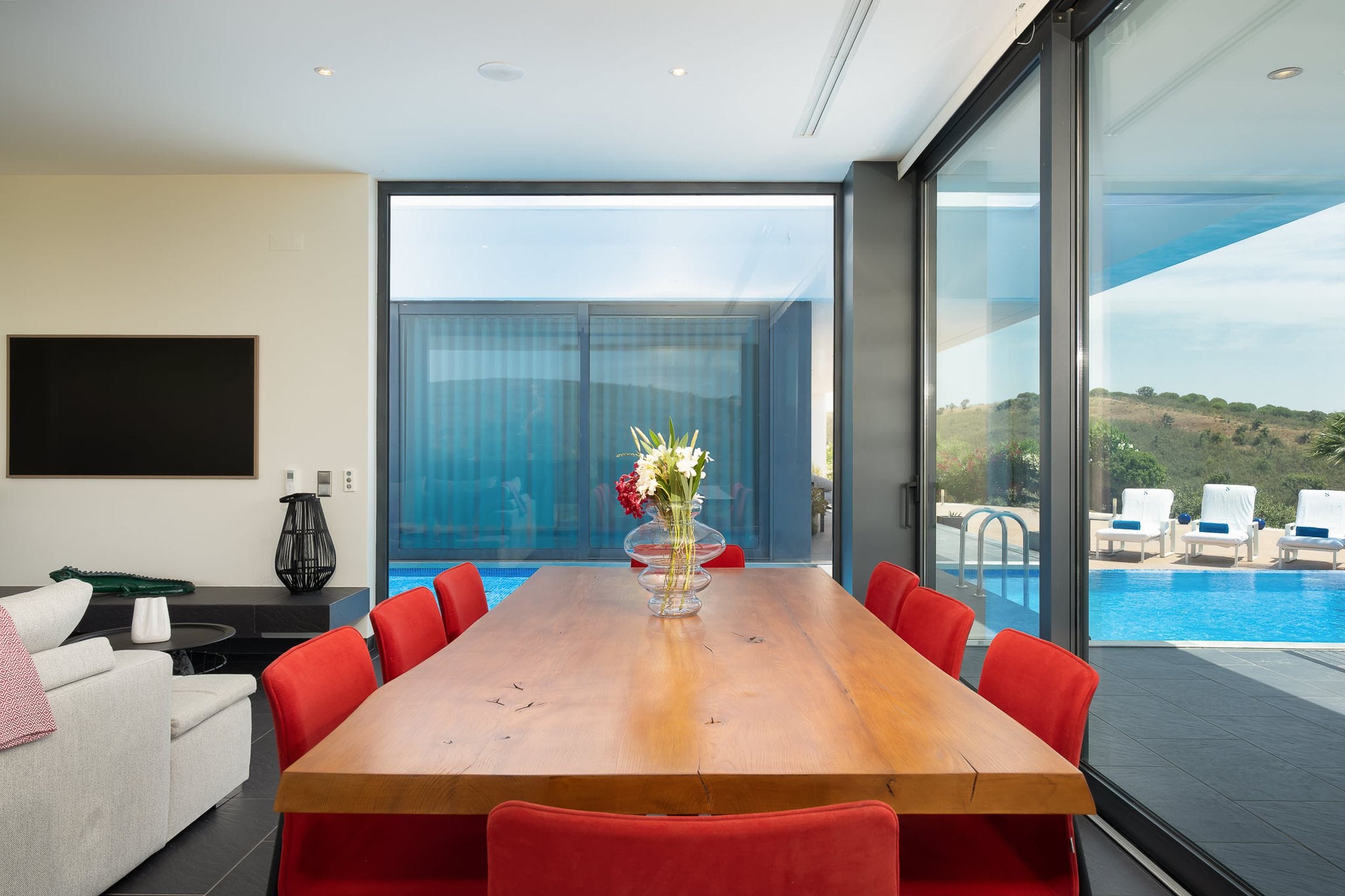 Live-edge timber dining table with red chairs beside glass walls overlooking the pool