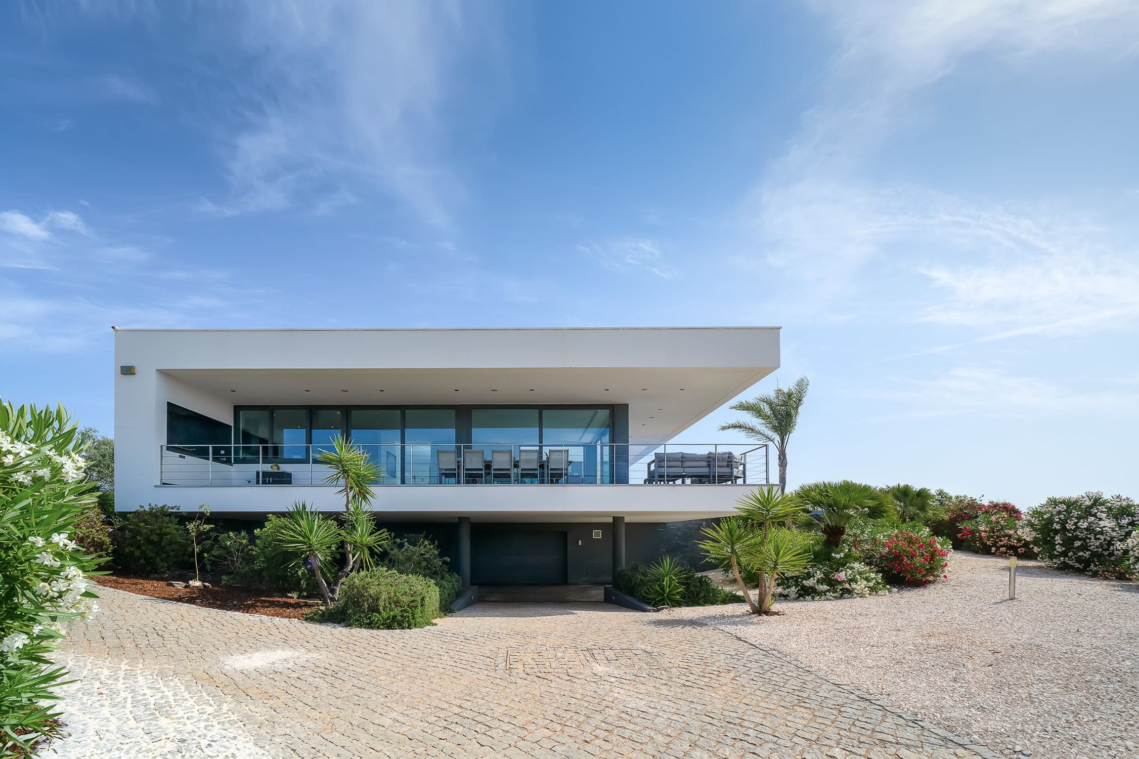 White cantilevered upper storey with a glass-lined terrace floating over a planted garden and cobbled drive