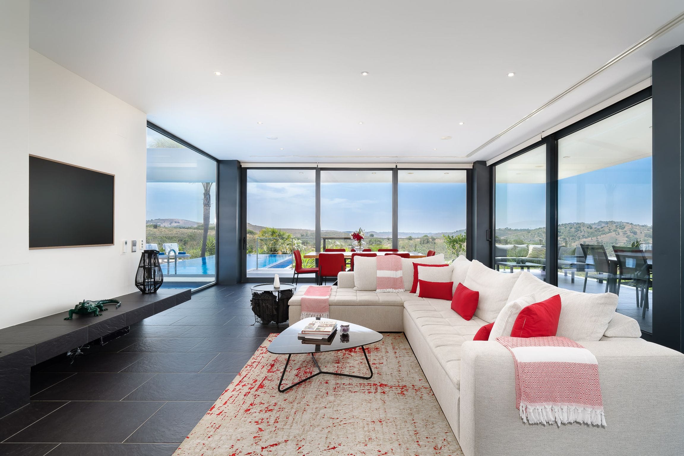 Open living room with a large white sectional sofa and red accents, glass walls opening to the pool and hills