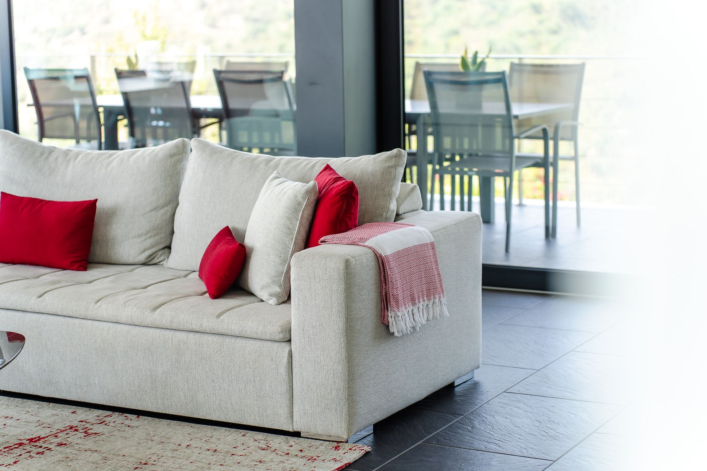 Soft white sofa with red cushions and a striped throw, terrace dining chairs beyond the glass