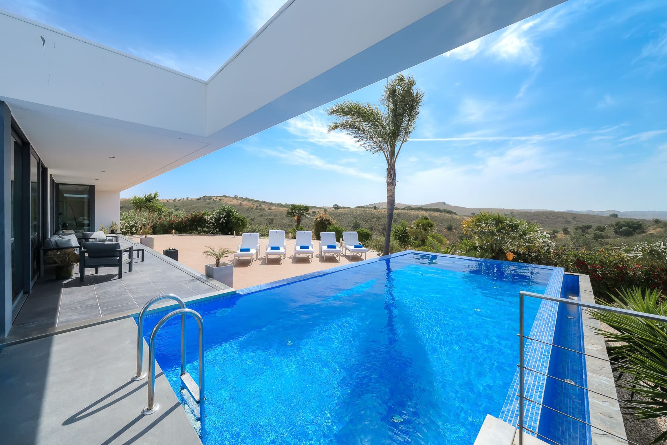 Infinity pool seen from beneath the villa's deep roof overhang, loungers and hills in the distance