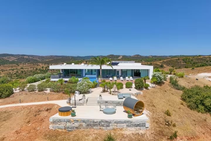 Aerial view of the villa's lower spa terrace with a barrel sauna, hot tub and fire pit on a stone-walled deck, hills beyond